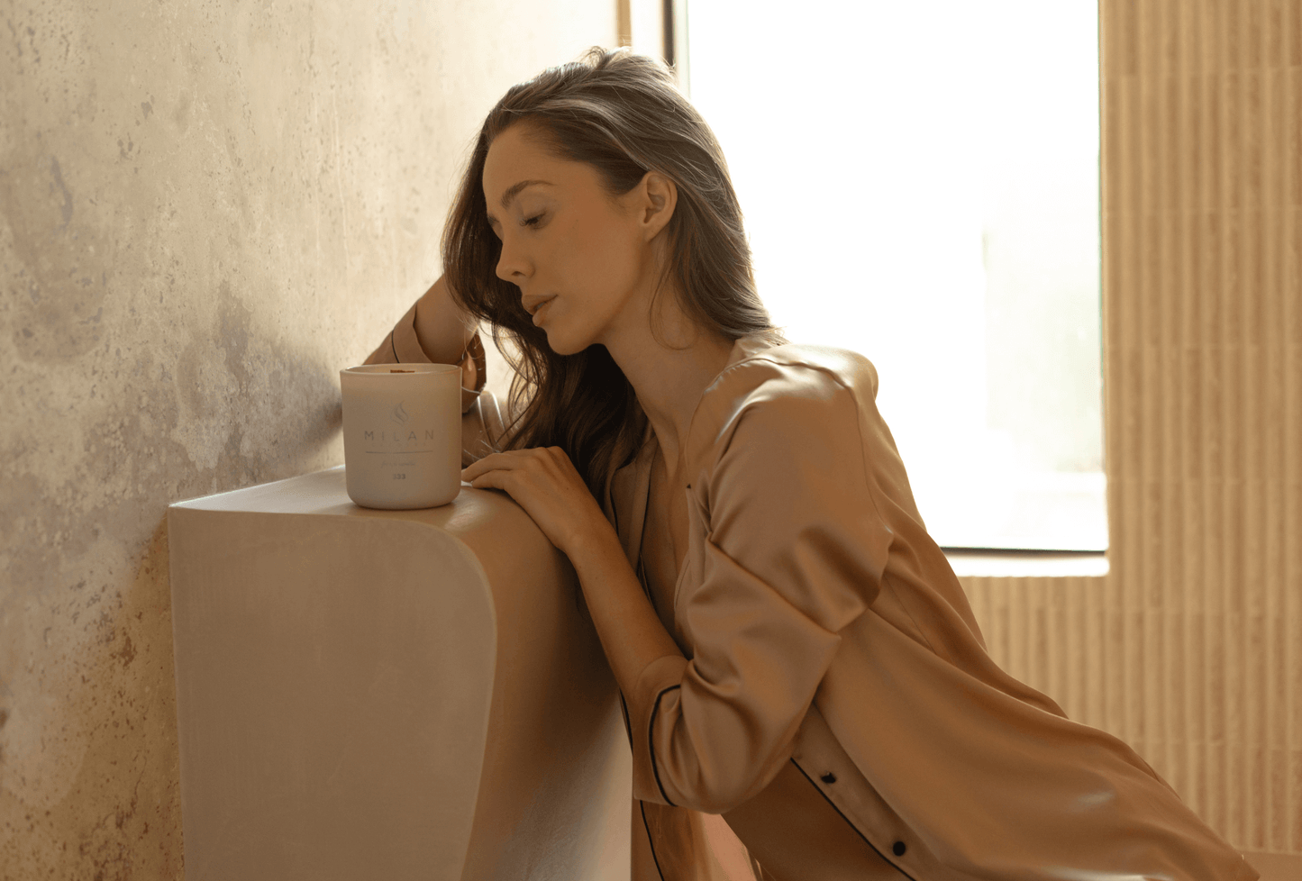 Woman in a natural loungewear leaning against a wall with a Milan Candle in a softly lit room.