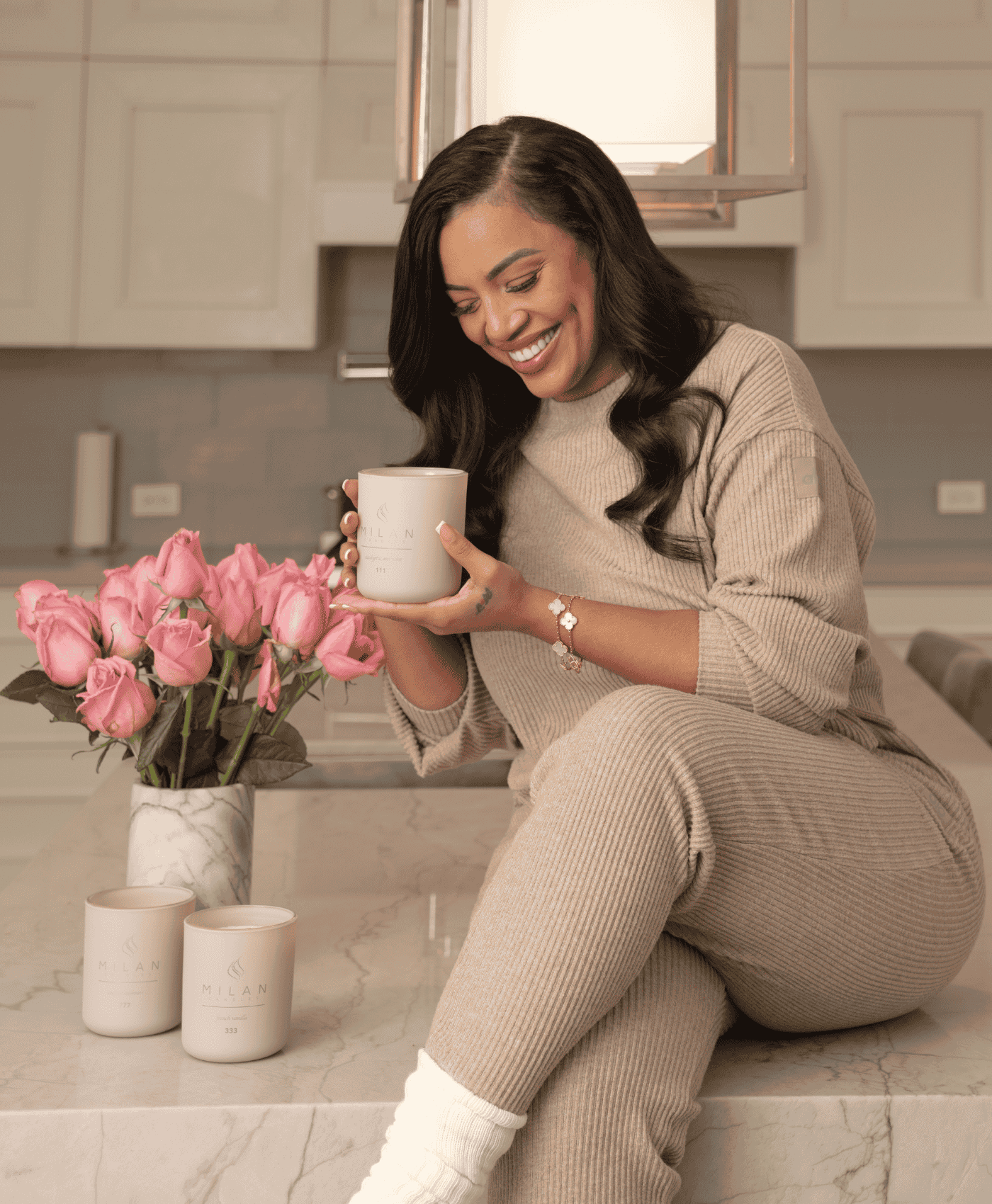 Woman sitting on a kitchen counter holding a luxury Milan Candle, smiling.